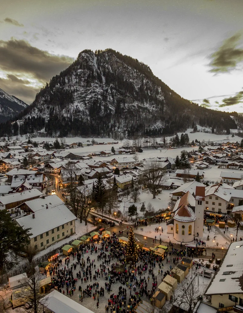 Blick von oben auf den beleuchteten Weihnachtsmarkt und den winterlichen Ortskern von Pfronten.