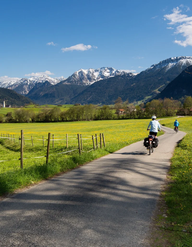 Radfahrer auf einem Radweg mit frühlingshaftem Blick auf grüne Wiesen und weiße Berggipfel.