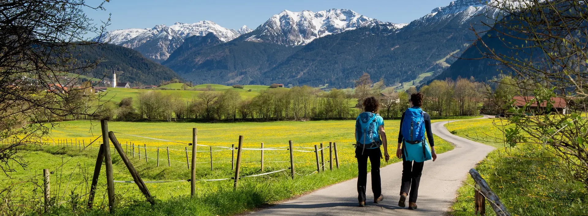 Zwei Wanderer auf einem schönen Weg durch das frühlingshafte Pfrontener Tal mit Blick auf die weißen Berggipfel.