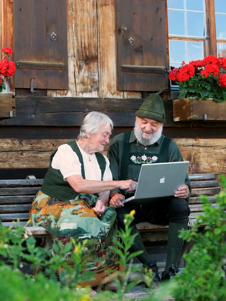 Zwei Personen in Tracht sitzen auf einer Bank und schauen auf ihren Laptop.