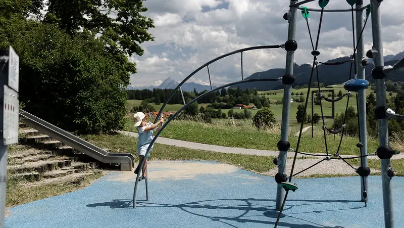 Ein Mädchen spielt auf einem Klettergerüst, im Hintergrund ist eine Hügellandschaft.