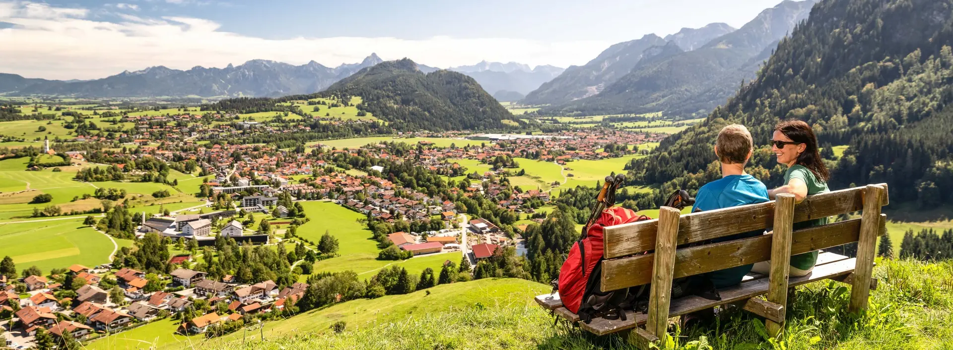 Zwei Wanderer sitzen auf einer Bank auf dem Edelsberg und genießen die Aussicht über Pfronten und die dahinterliegende Berglandschaft.