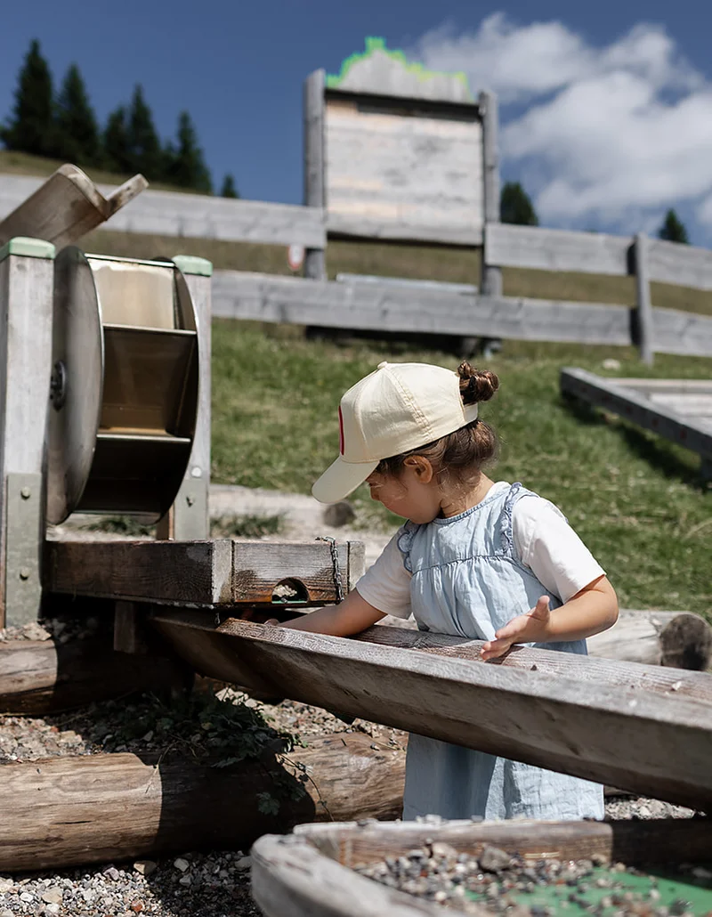 Ein kleines Mädchen spielt an einem hölzernen Wasserspielplatz auf einer Wiese bei sonnigem Wetter.