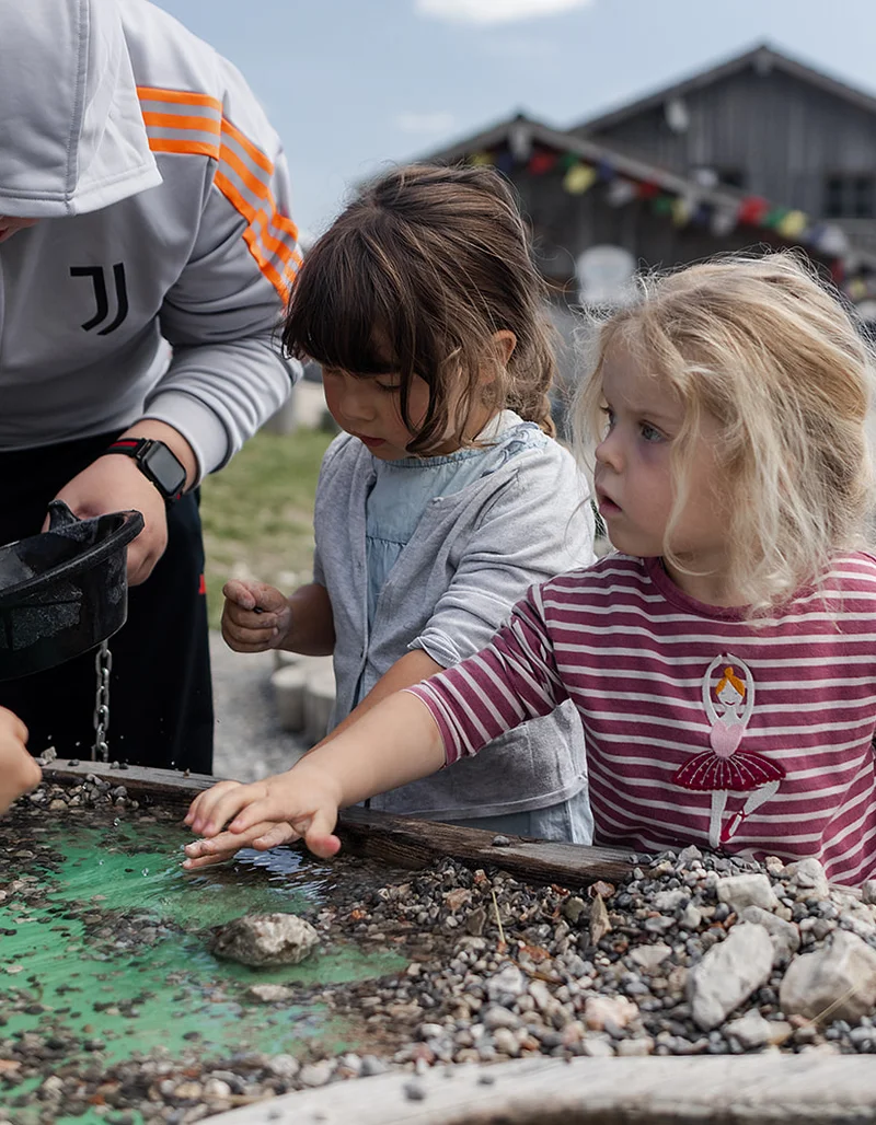 Zwei Kinder spielen an einem Wasserspieltisch mit Steinen und Kies, während ein Erwachsener beim „Schürfen“ hilft.
