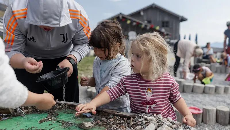 Zwei Kinder spielen an einem Wasserspieltisch mit Steinen und Kies, während ein Erwachsener beim „Schürfen“ hilft.