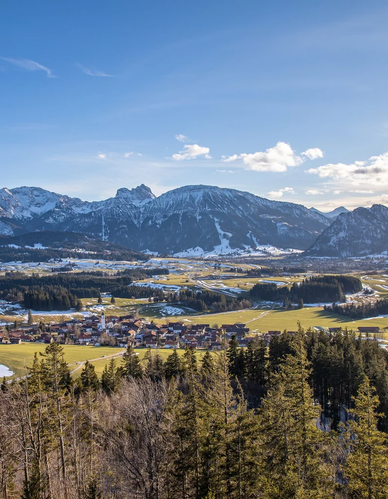 Panoramablick auf das frühlingshafte Pfronten und die schneebedeckte Bergwelt.