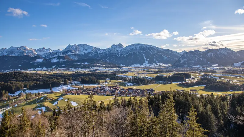 Panoramablick auf das frühlingshafte Pfronten und die schneebedeckte Bergwelt.