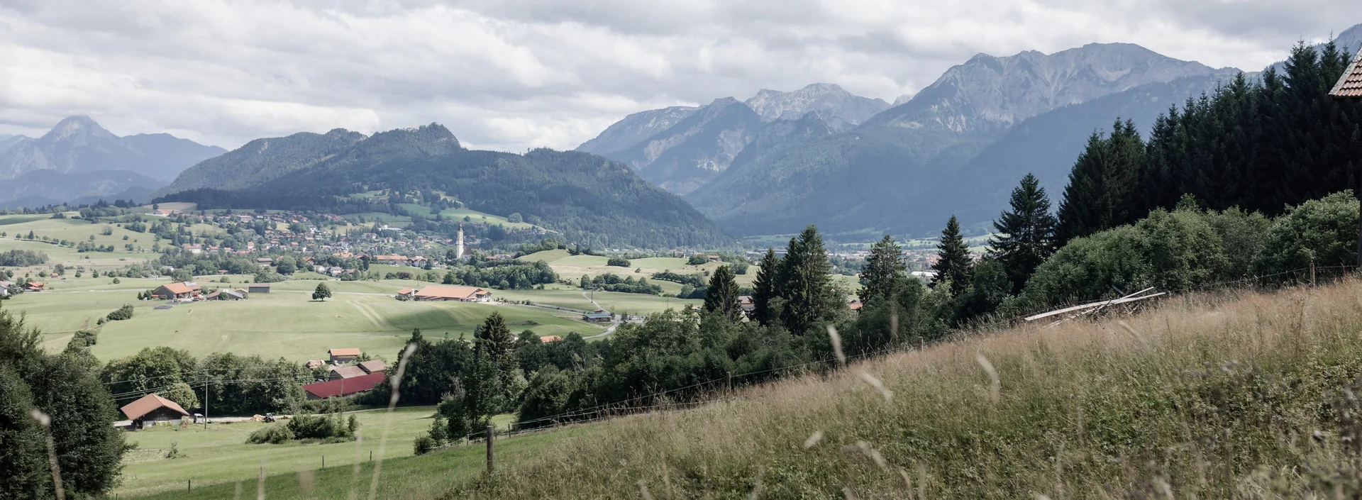 Alpenlandschaft mit Wiesen, Dorf und Bergen im Hintergrund.