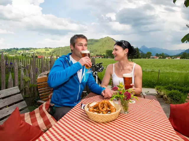 Essen im Biergarten in Pfronten im Allgäu Zwei Personen bei der Einkehr im Biergarten, im Hintergrund sind sommerlich grüne Wiesen zu sehen.
