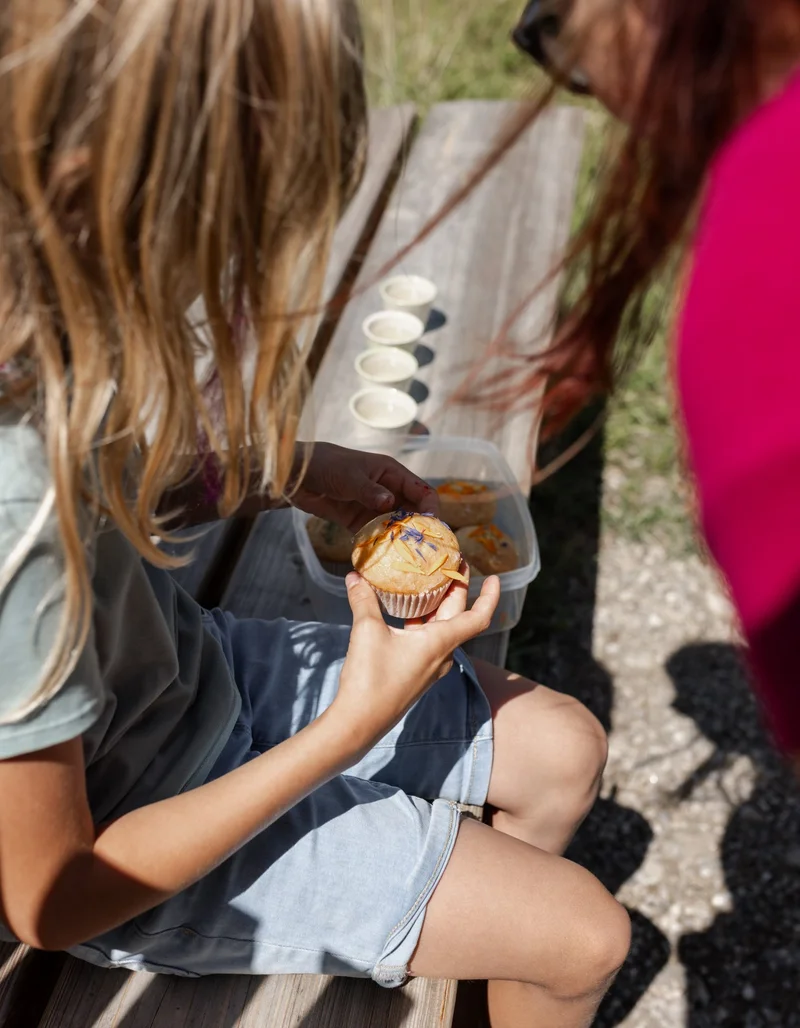 Kind sitzt auf einer Holzbank und hält einen Muffin mit essbaren Blüten in der Hand.