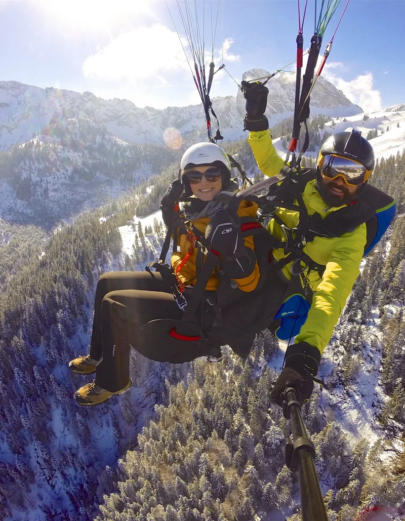 Ein Tandemflug mit dem Gleitschirm bei Sonnenschein mit Blick auf eine winterliche Berglandschaft.