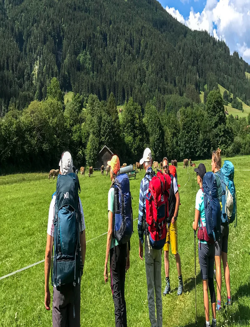 Eine Gruppe von Wanderern mit bepackten Rucksäcken steht auf einer grünen Wiese und blickt in Richtung der Berge.