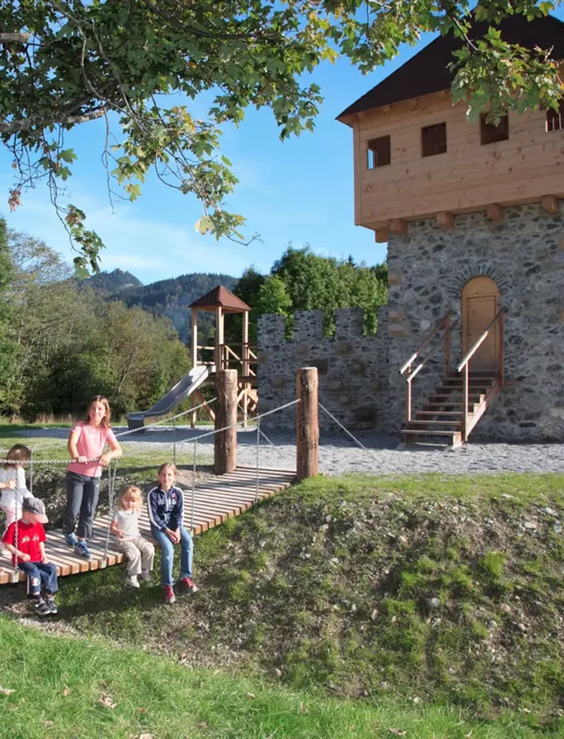 Kinder stehen auf einer kleinen Hängebrücke, im Hintergrund ist die nachgebaute Burgenanlage auf dem Ritterspielplatz in Pfronten zu sehen.
