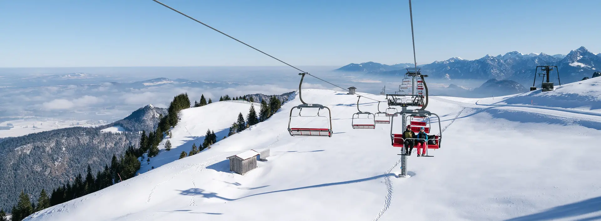 Blick von der Sesselbahn am Breitenberg über eine idyllisch verschneite Winterlandschaft mit den Allgäuer Bergen im Hintergrund.