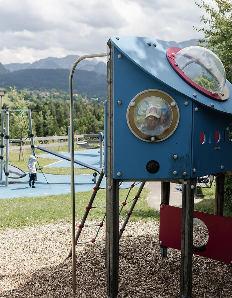 Ein Kind schaut aus einem Bullauge eines raketenförmigen Spielgeräts auf dem Planetenspielplatz, im Hintergrund sind Kletterelemente und eine grüne Hügellandschaft mit Bergen.
