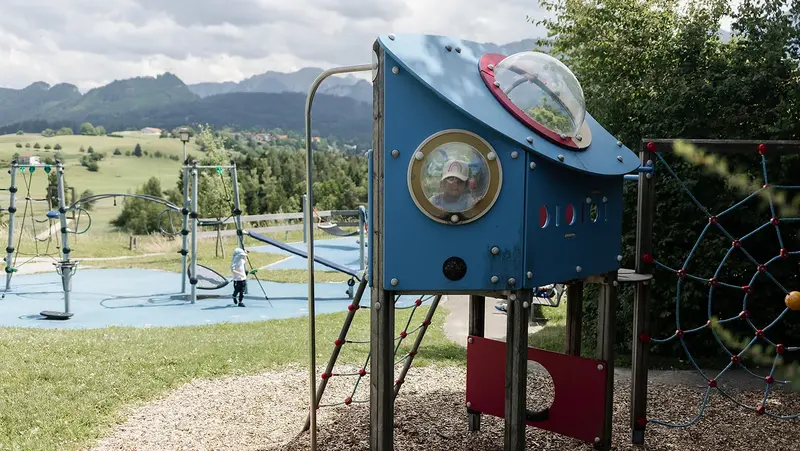 Ein Kind schaut aus einem Bullauge eines raketenförmigen Spielgeräts auf dem Planetenspielplatz, im Hintergrund sind Kletterelemente und eine grüne Hügellandschaft mit Bergen.