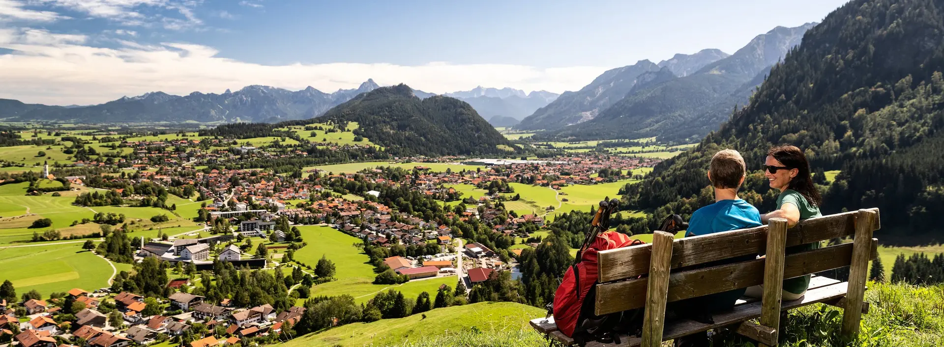 Zwei Wanderer sitzen auf einer Bank auf dem Edelsberg und genießen die Aussicht über Pfronten und die dahinterliegende Berglandschaft.