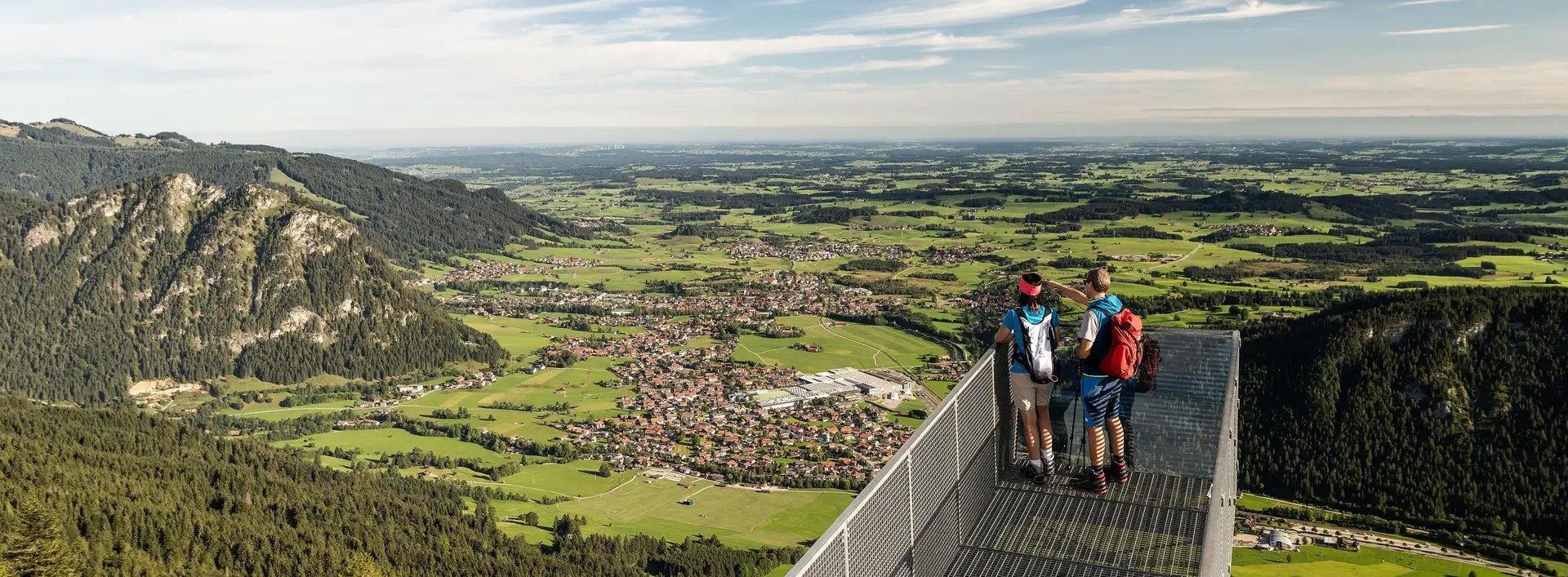 Zwei Wanderer auf dem Aussichtssteg am Breitenberg mit weitem Blick in das Pfrontener Tal.
