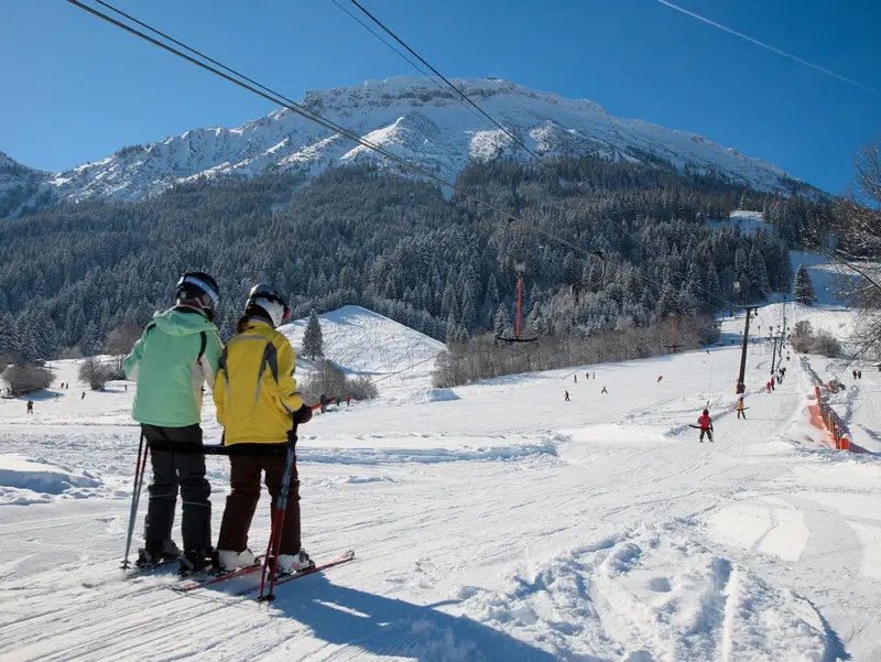Skifahrer werden im Skilift auf den Berg befördert, im Hintergrund sind Skipisten und Berge zu sehen.