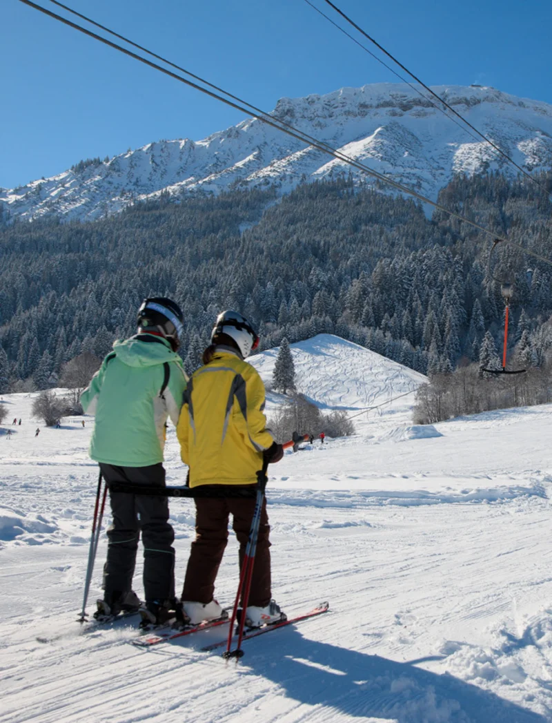 Skifahrer werden im Skilift auf den Berg befördert, im Hintergrund sind Skipisten und Berge zu sehen.