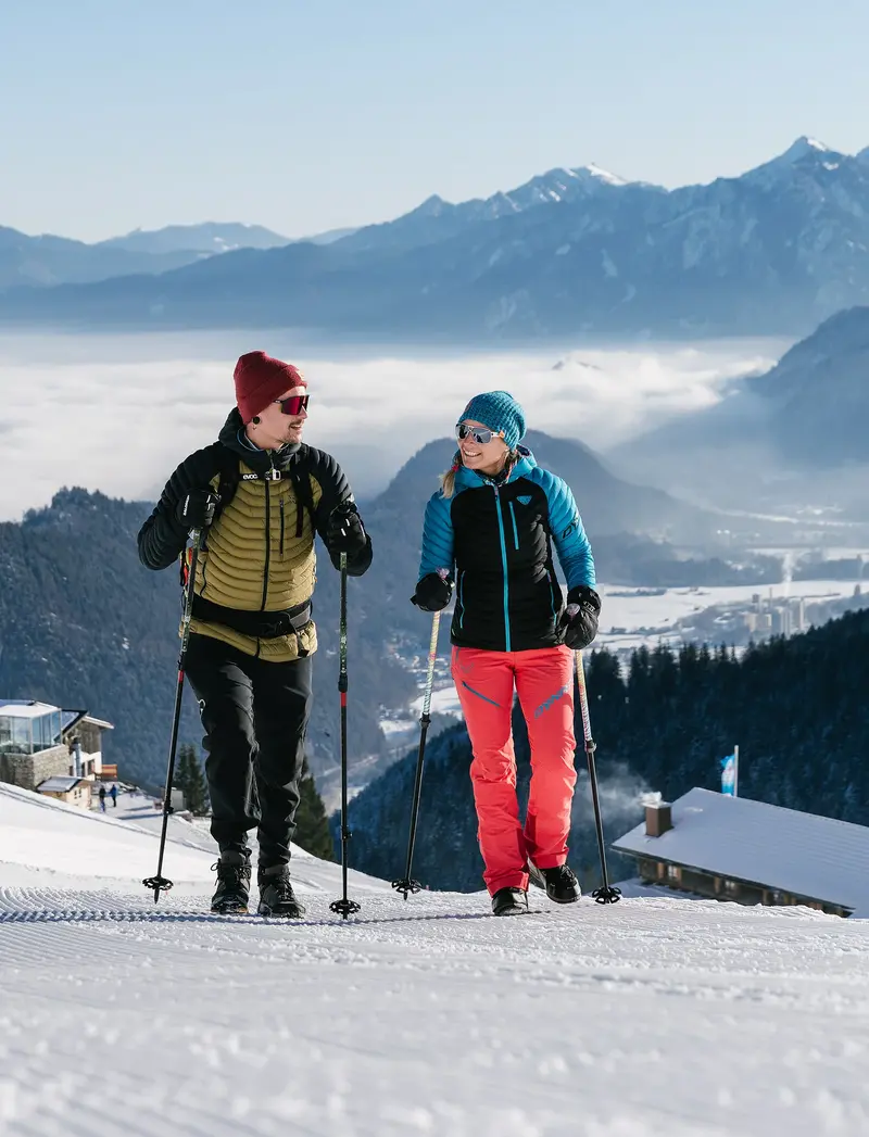 Zwei Winterwanderer auf präparierten Wegen, im Hintergrund eine verschneite Berglandschaft.