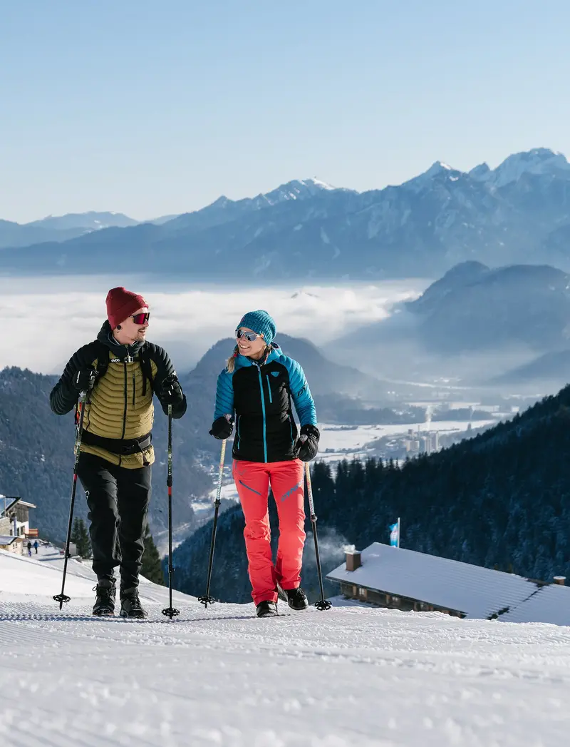 Zwei Winterwanderer auf präparierten Wegen, im Hintergrund eine verschneite Berglandschaft.