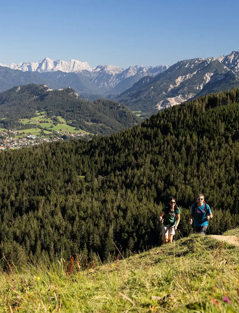 Zwei Wanderer auf einem schmalen Pfad auf dem Berg inmitten grüner Wiesen und einer beeindruckenden Bergkulisse.