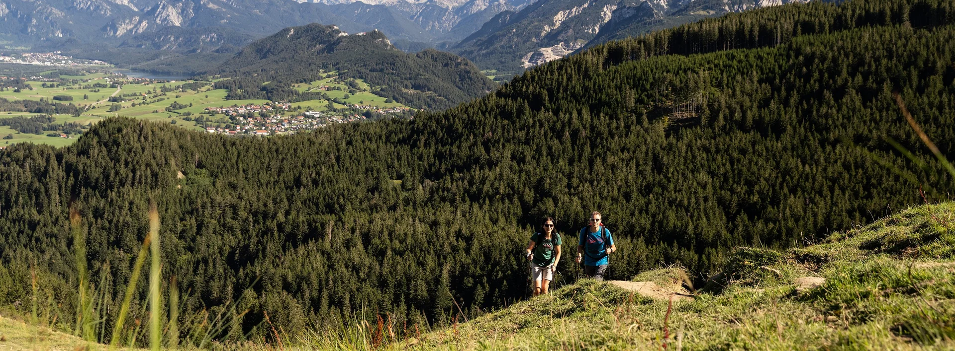 Zwei Wanderer auf einem schmalen Pfad auf dem Berg inmitten grüner Wiesen und einer beeindruckenden Bergkulisse.