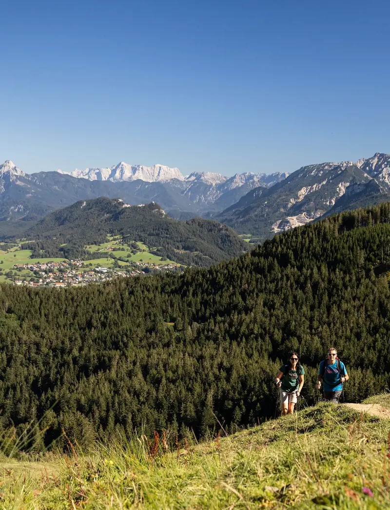 Zwei Wanderer auf einem schmalen Pfad auf dem Berg inmitten grüner Wiesen und einer beeindruckenden Bergkulisse.