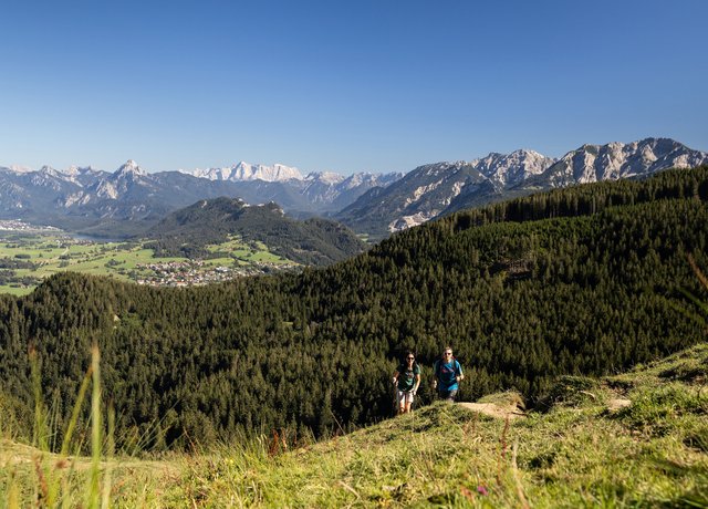 Zwei Wanderer auf einem schmalen Pfad auf dem Berg inmitten grüner Wiesen und einer beeindruckenden Bergkulisse.