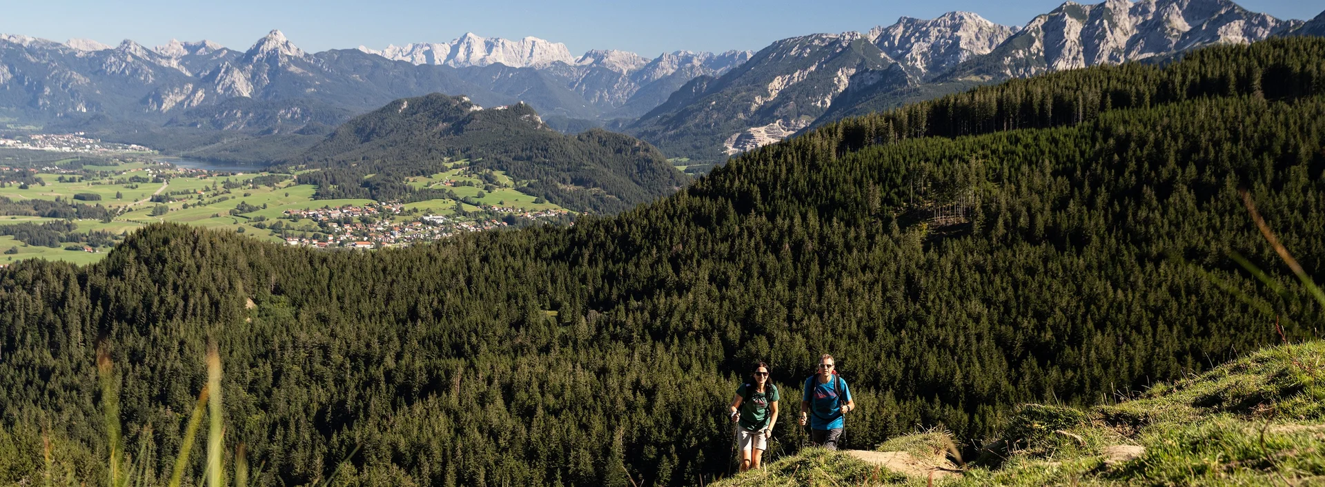 Zwei Wanderer auf einem schmalen Pfad auf dem Berg inmitten grüner Wiesen und einer beeindruckenden Bergkulisse.