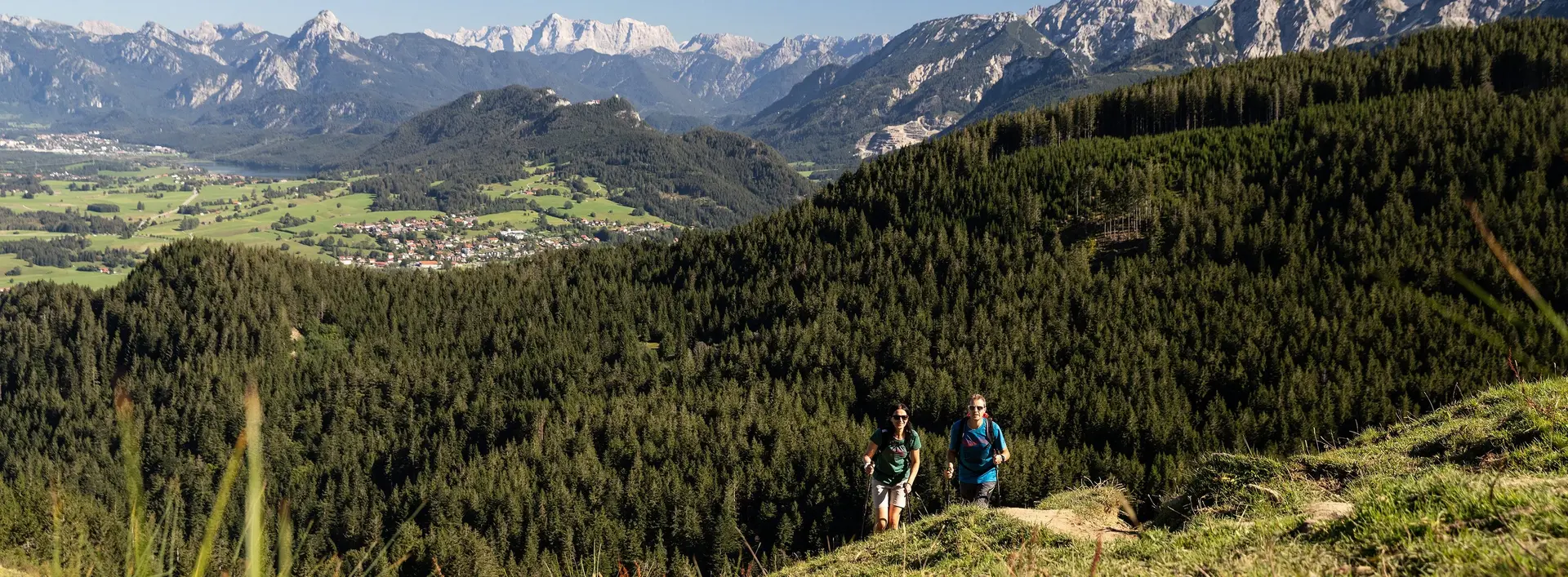 Zwei Wanderer auf einem schmalen Pfad auf dem Berg inmitten grüner Wiesen und einer beeindruckenden Bergkulisse.