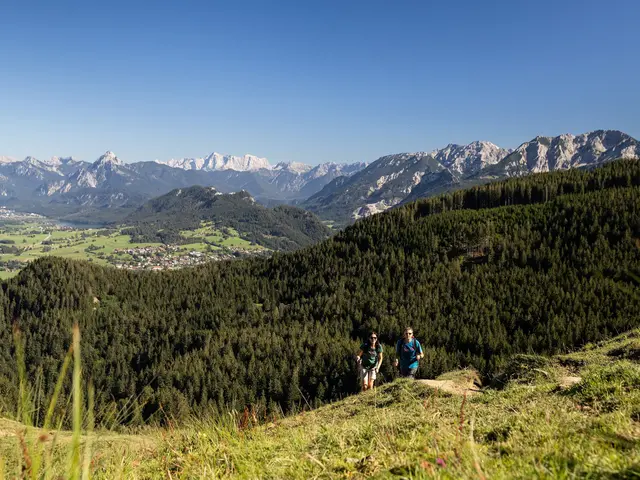 Zwei Wanderer auf einem schmalen Pfad auf dem Berg inmitten grüner Wiesen und einer beeindruckenden Bergkulisse.