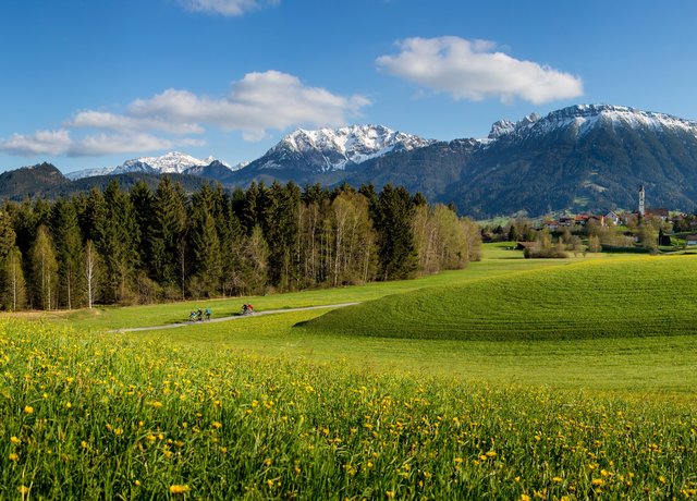 Frühlingshafter Panoramablick über die Wiesen Pfrontens mit weißen Berggipfeln.