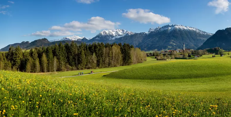 Frühlingshafter Panoramablick über die Wiesen Pfrontens mit weißen Berggipfeln.