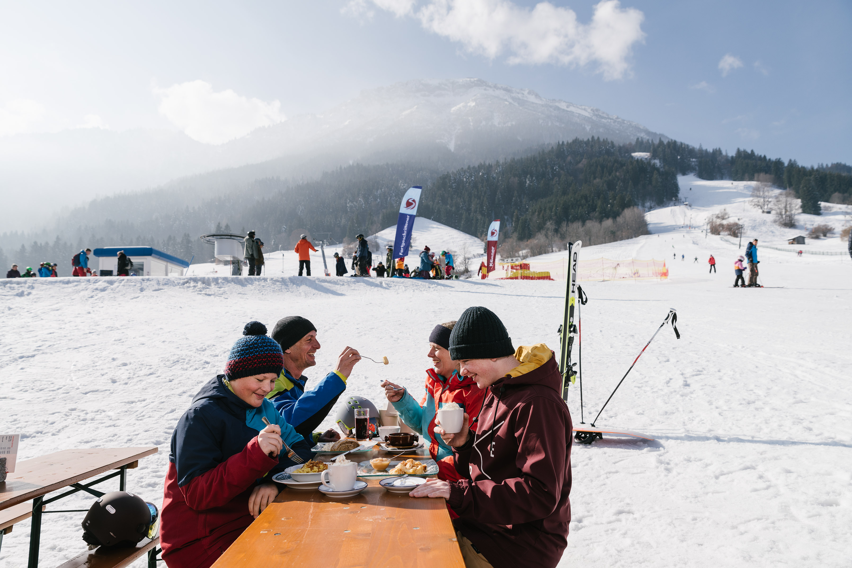 Eine Familie bei der Einkehr an der Skipiste, im Hintergrund eine verschneite Landschaft und Skipisten.