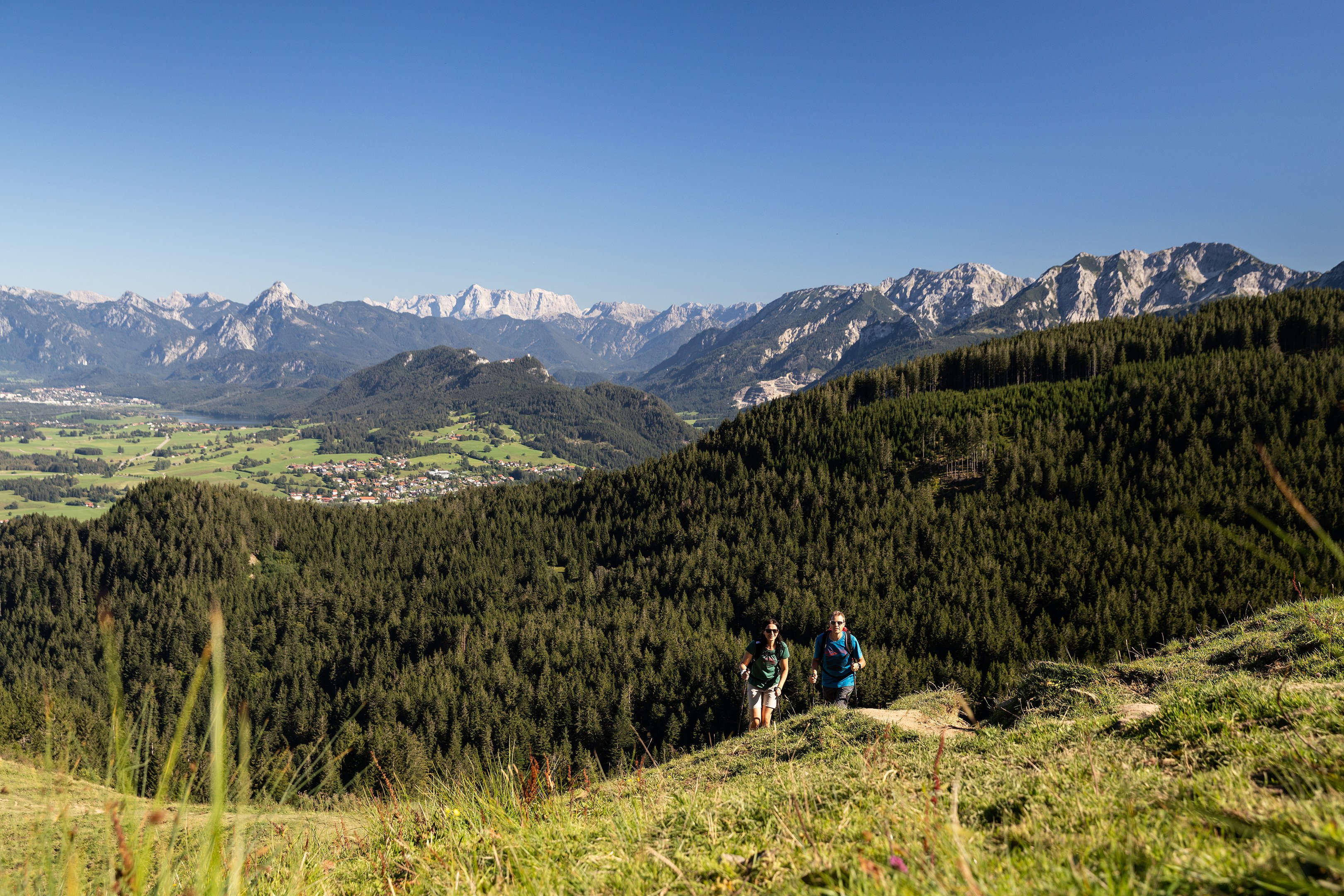 Zwei Wanderer auf einem schmalen Pfad auf dem Berg inmitten grüner Wiesen und einer beeindruckenden Bergkulisse.